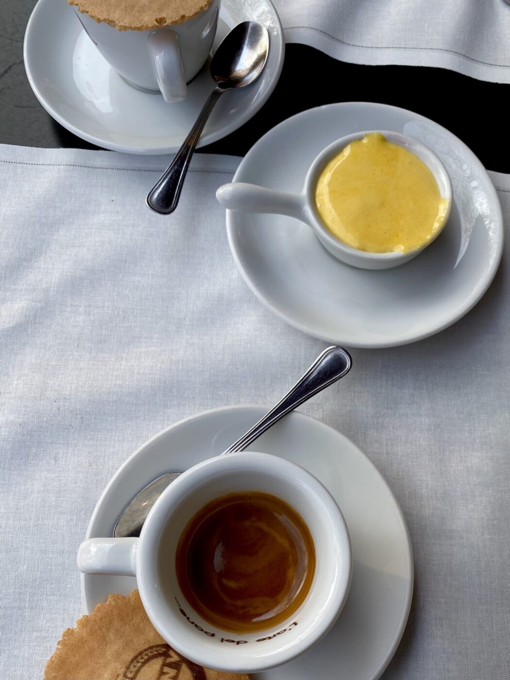 an overhead photo of a cup of espresso, cup of yellow, thick zabaione, and another cup of espresso half in the frame all on saucers and on a white tablecloth