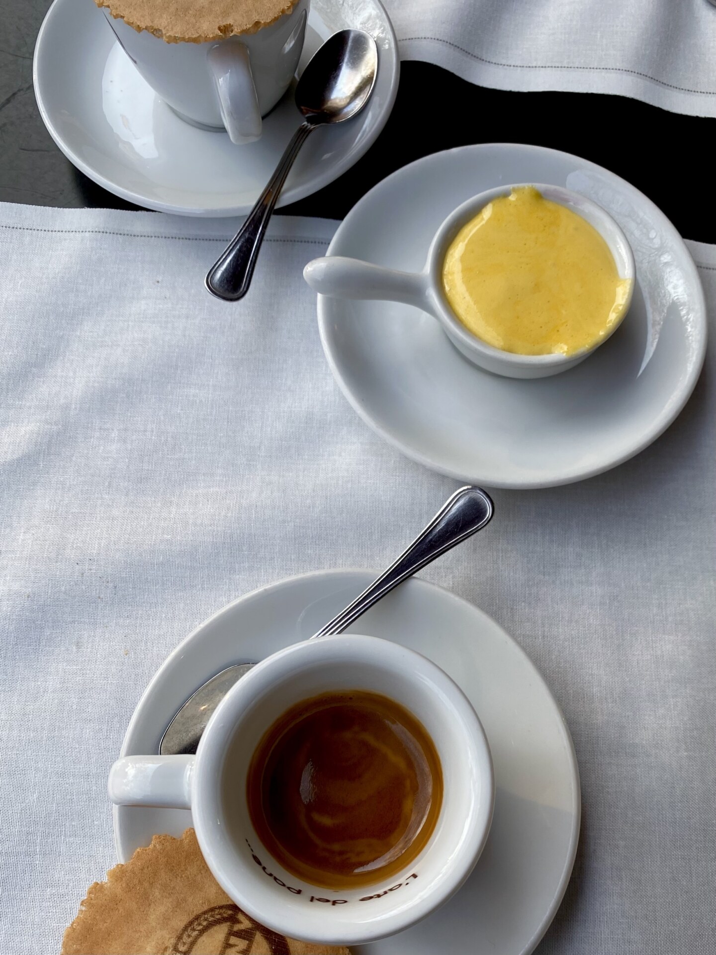 an overhead photo of a cup of espresso, cup of yellow, thick zabaione, and another cup of espresso half in the frame all on saucers and on a white tablecloth