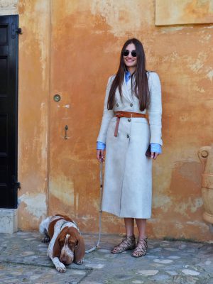 Margherita Cardelli smiling wearing clear overcoat and sunglasses in front of a antique orange wall and a dog laid down