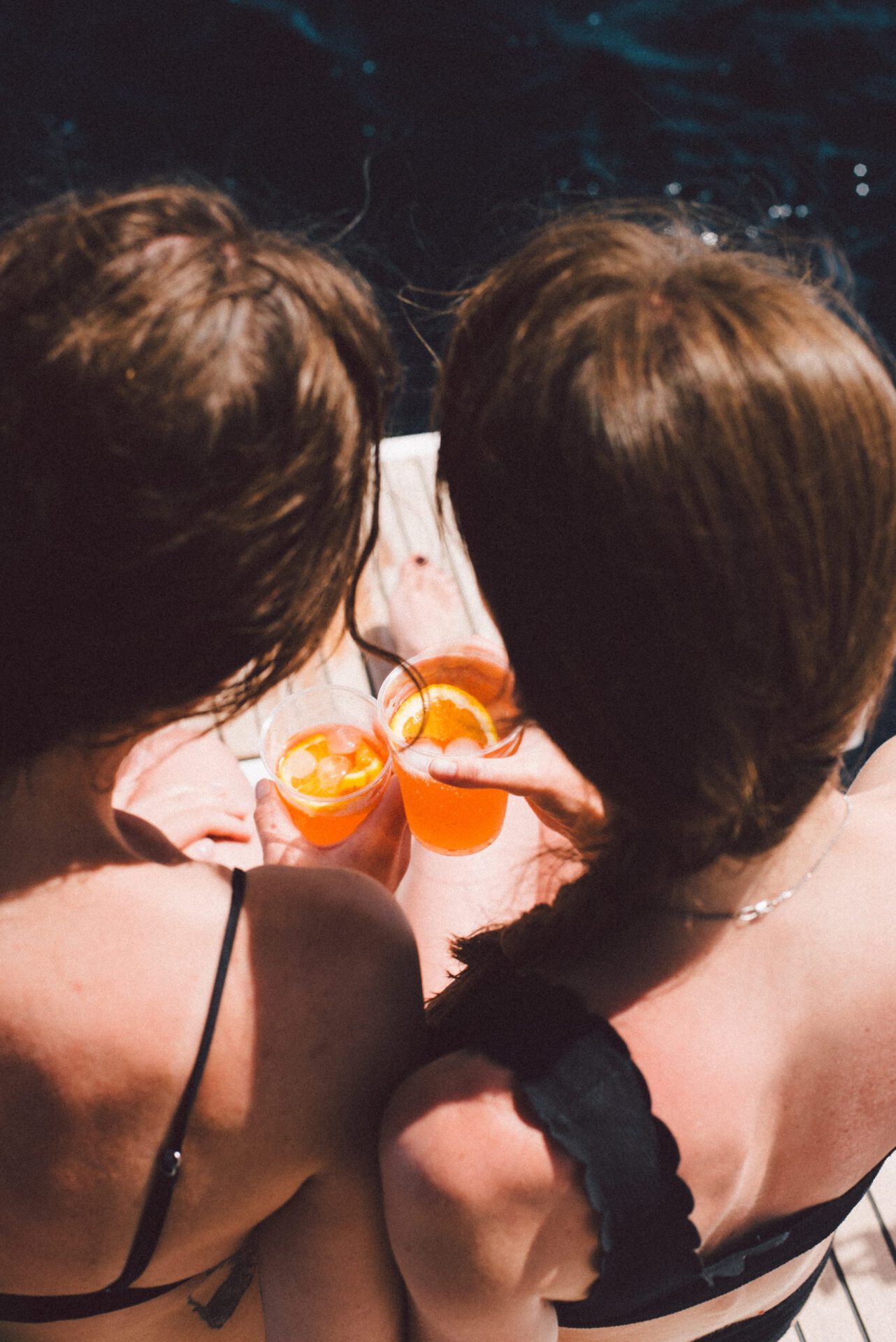 Two women facing back holding aperol spritz cocktail cups over a reck on the sea Hand palm facing to a mountain sea and clouds in Panarea of the Aeolian Islands in Sicily Italy