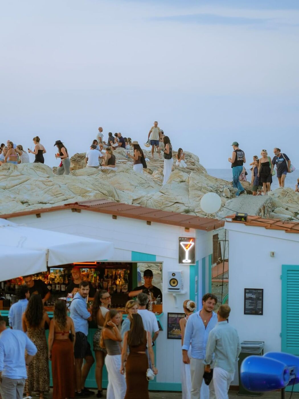 People gather on rocky cliffs and at a turquoise beach bar under a clear sky, chatting, ordering drinks, or admiring the coastal view.
