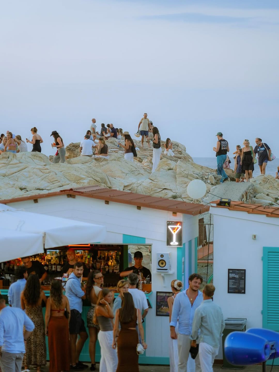 People gather on rocky cliffs and at a turquoise beach bar under a clear sky, chatting, ordering drinks, or admiring the coastal view.