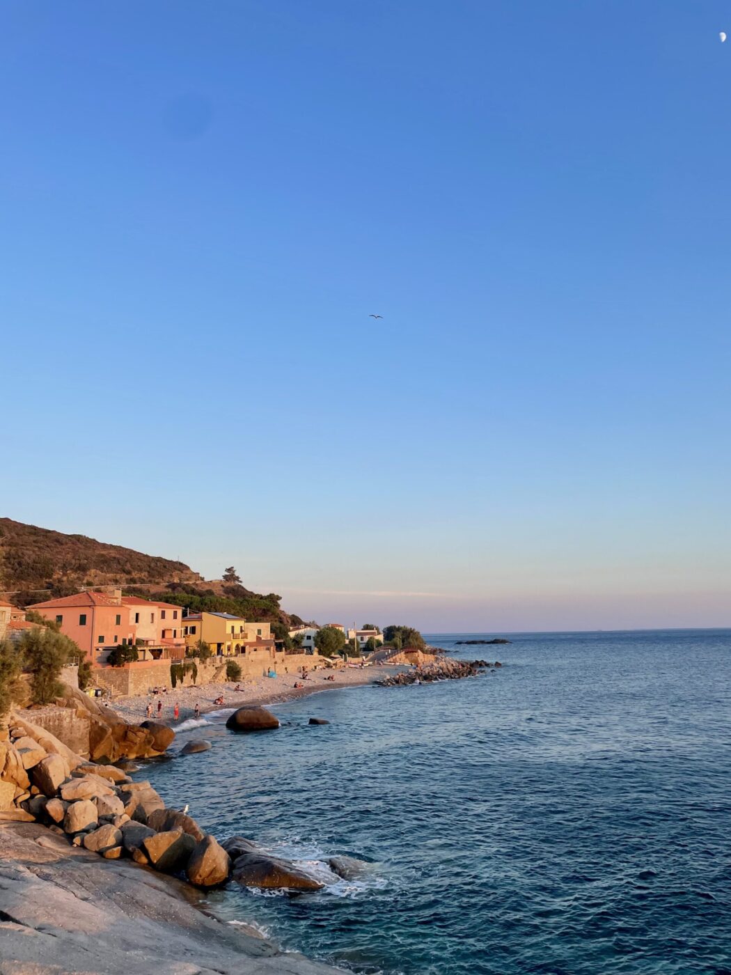 Colorful houses along a rocky shore at sunset, people stroll on the beach, calm blue sea, clear sky, and hills in the distance.