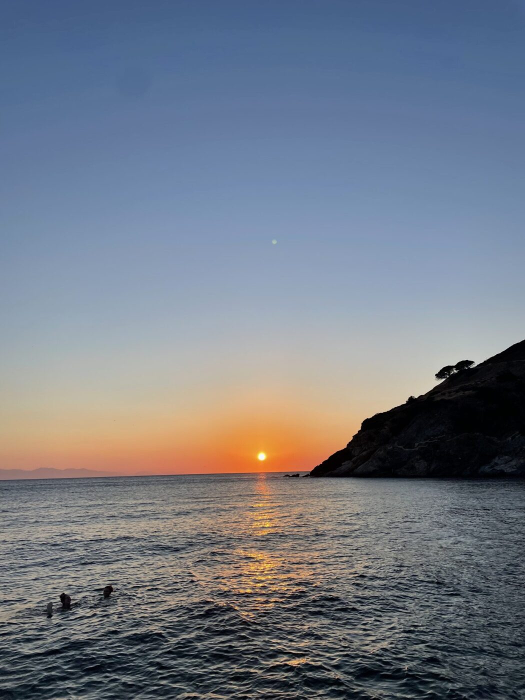 The sun sets over a calm sea, orange reflecting on water; silhouetted hill and trees right, two people swim, sky fades blue to orange.