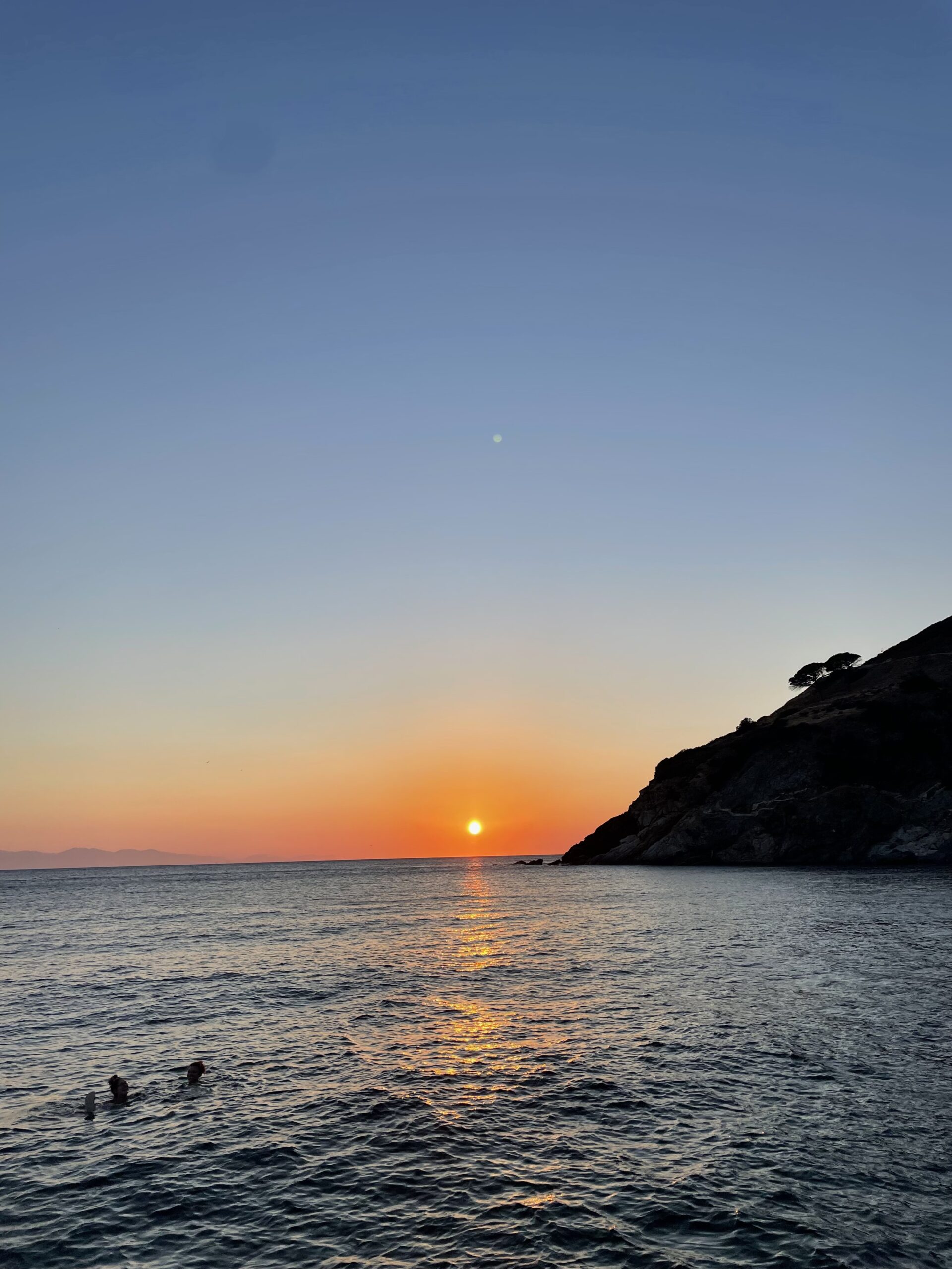 The sun sets over a calm sea, orange reflecting on water; silhouetted hill and trees right, two people swim, sky fades blue to orange.