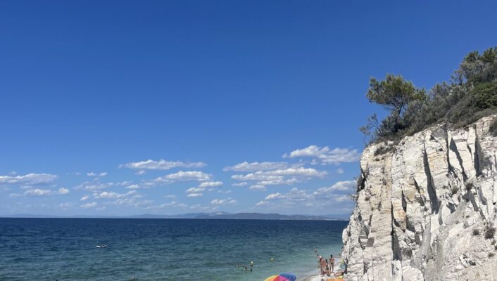 Le persone si rilassano sotto gli ombrelloni luminosi su una spiaggia di ciottoli vicino all'acqua turchese e alle scogliere bianche, con il cielo azzurro e la terra lontana.