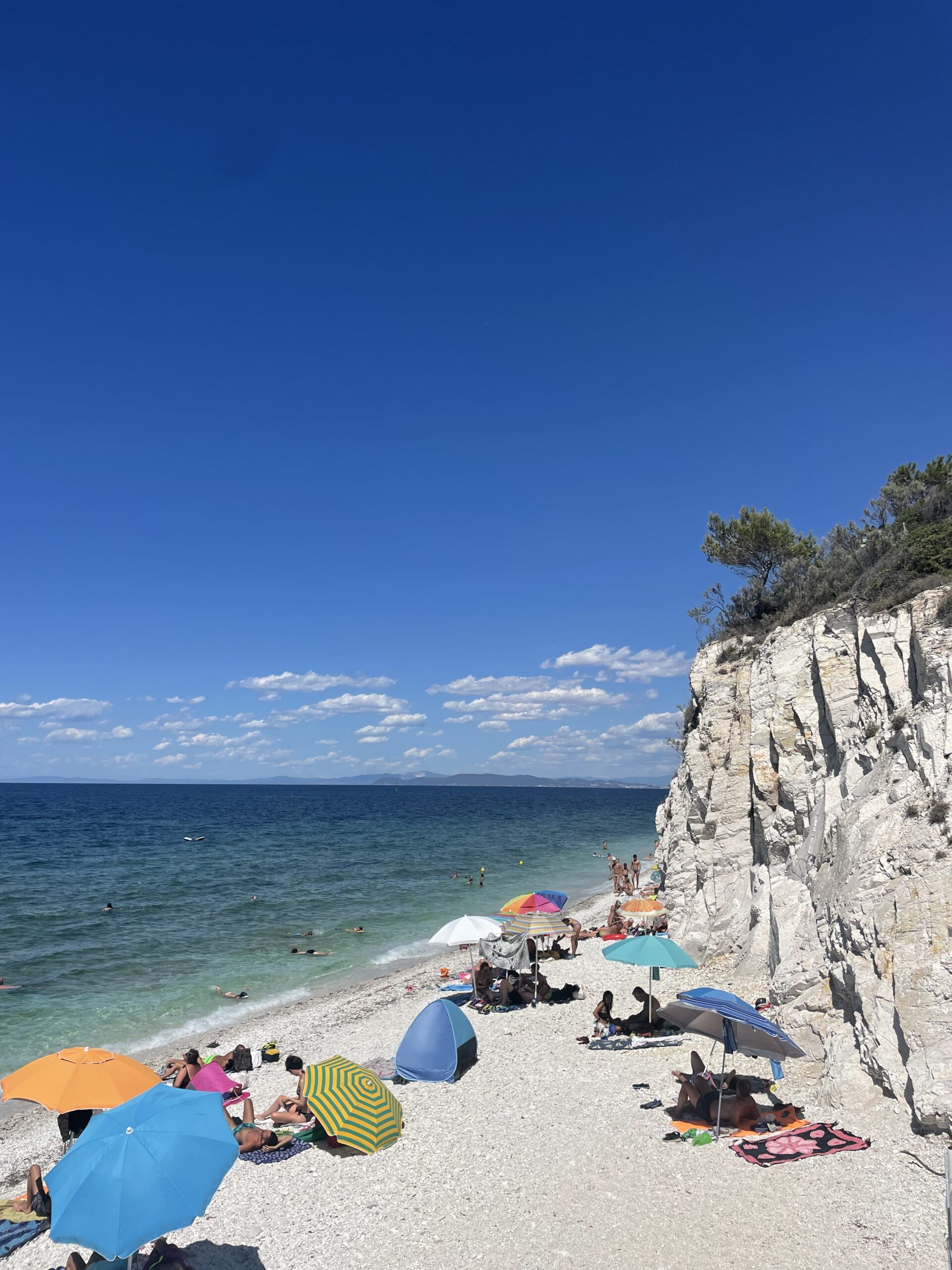 People lounge under bright umbrellas on a rocky beach near white cliffs, turquoise sea, swimmers, and trees atop the cliff under blue sky.