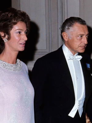 Angelli at a Formal Event - Angelli, in a light pink dress with jeweled details, stands beside a man in a black tuxedo with a white bow tie and medals. They appear to be attending a formal event in an elegant setting.