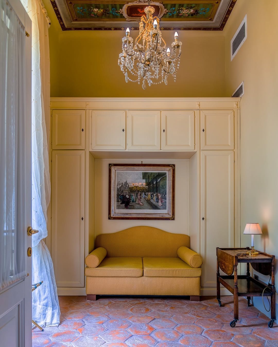 A cozy room with yellow walls, cabinets, and sofa; a painting overhead, chandelier, side table lamp, tile floor, and painted ceiling.