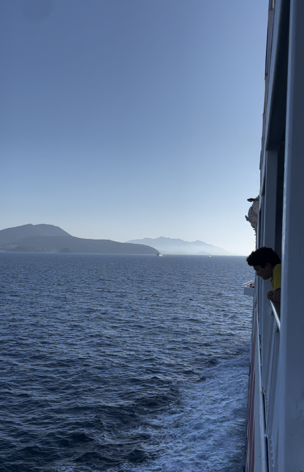A person leans on a ship’s railing, gazing at the sea; distant mountains and hazy sky, with the ship’s wake trailing behind.