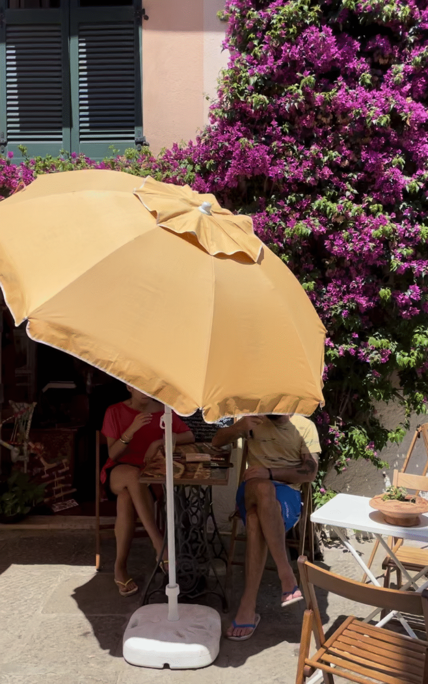 Two people at an outdoor café under a yellow umbrella beside a wall of purple flowers, with food and drinks and dappled sunlight.
