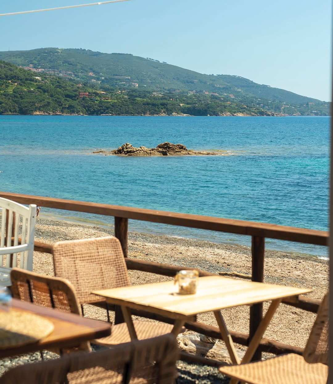 A seaside café with wicker chairs, wooden tables, and views of a calm blue sea, distant green hills, and a rocky outcrop nearby.