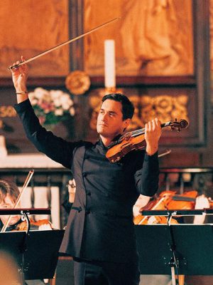 Charlie Siem formal dressed with dark clothes playing the violin with an orchestra in the background