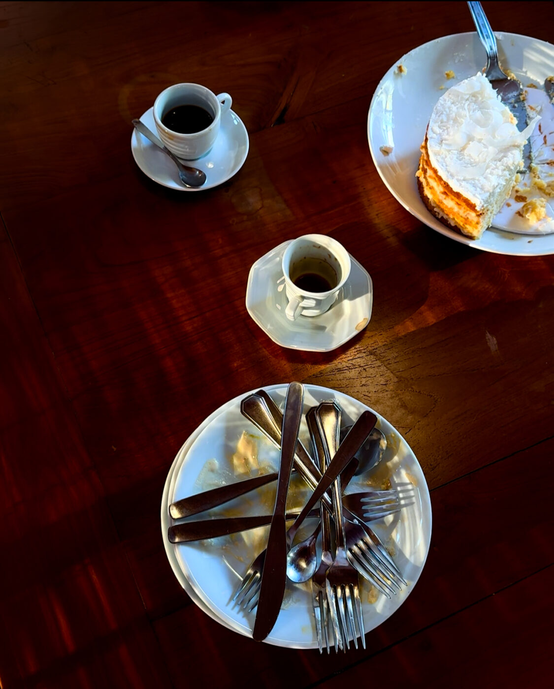wooden table with two coffee cups, a white coconut cake and a plate filled with dirty cutlery