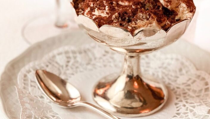 Tiramisu dessert dusted with cocoa powder over golden cup and white plate with spoon and glass of wine in the background in Italy