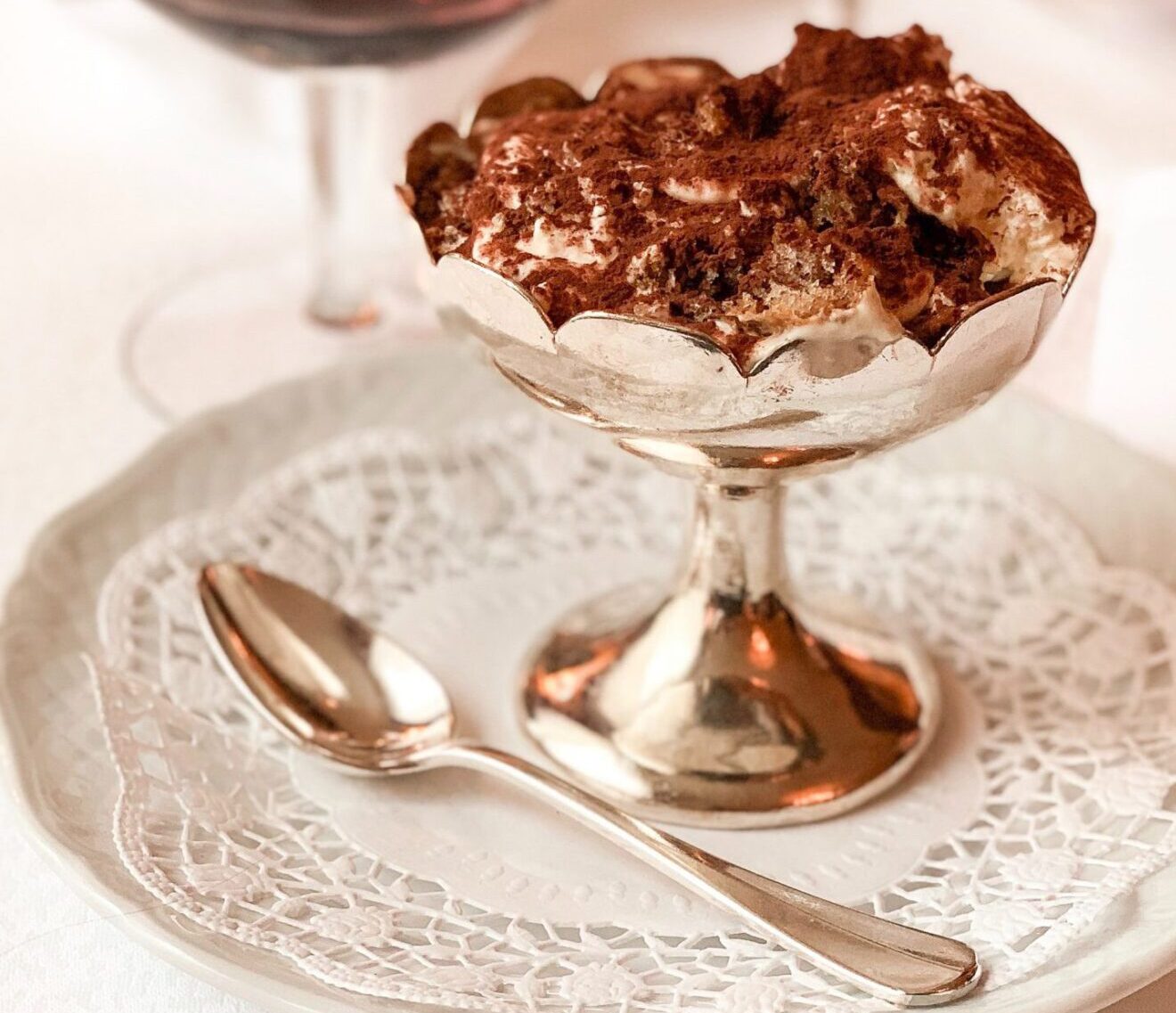 Tiramisu dessert dusted with cocoa powder over golden cup and white plate with spoon and glass of wine in the background in Italy