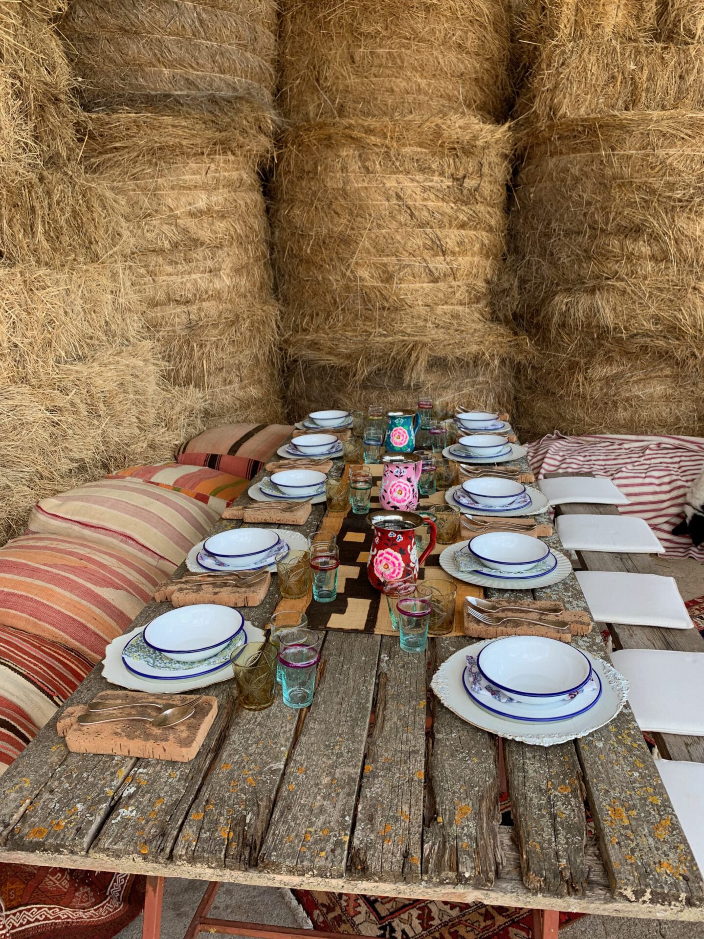 Wooden table with colorful ceramic plates and cups with pillows surrounded by hay in an equestrian farm, Tenuta di Paternostro in Tuscia Italy