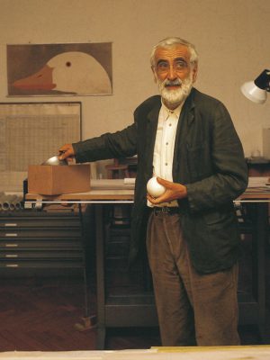 Enzo Mari formal dressed with glasses and holding an egg in his hand on an artistic studio filled up with tools plans and shelves in Italy