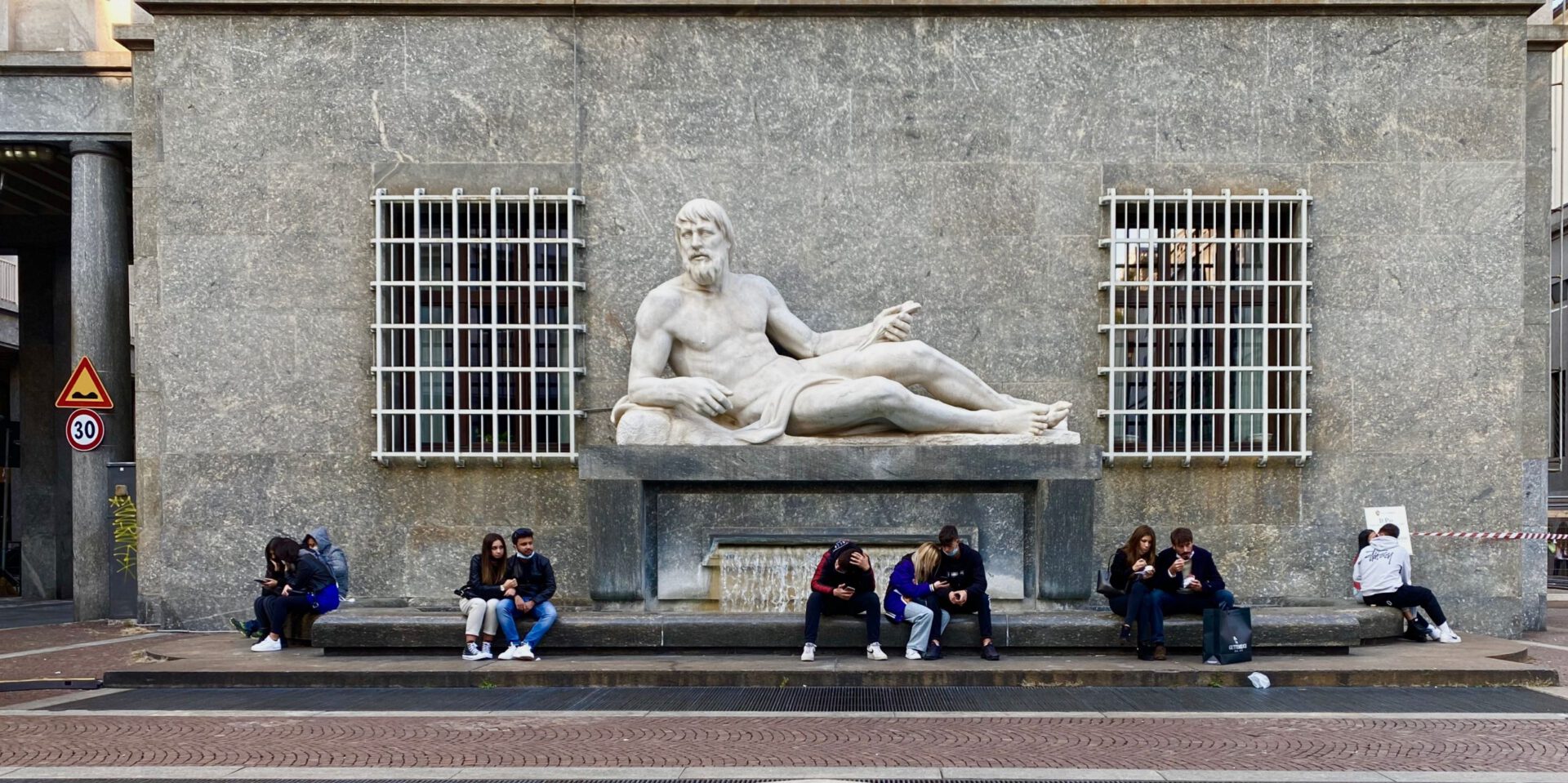 People seated on the floor in front of marble sculpture and grey wall at Piazza C.L.N. in Turin Piedmont in Italy