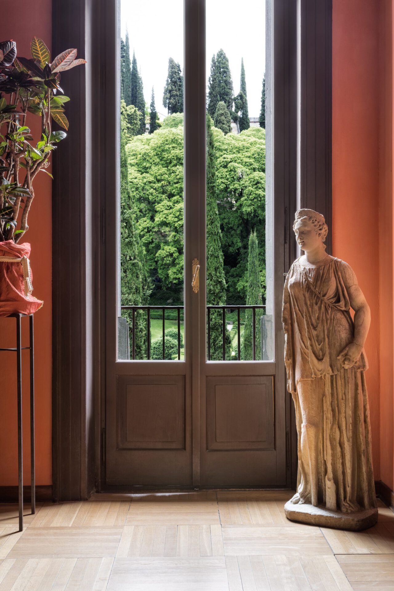 Wooden window viewing a park with trees and bushes in a warm orange room with sculptures at the Giardino Giusti in Veneto Italy
