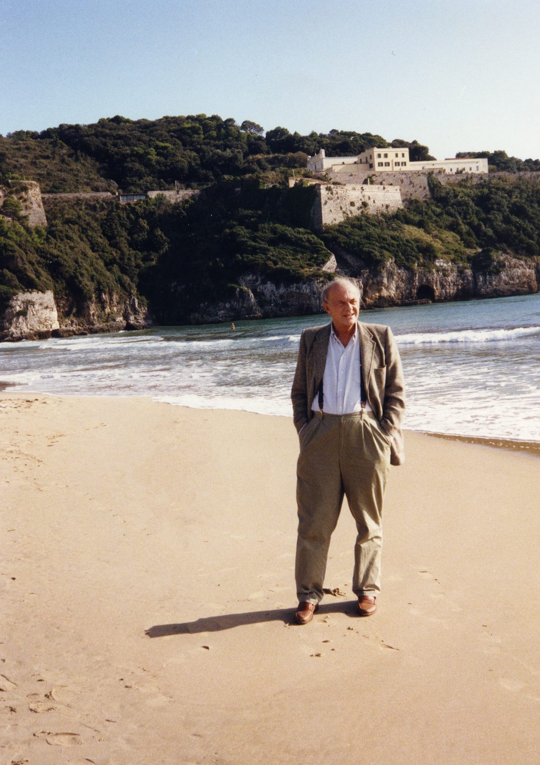 Cy Twombly formal dressed with brown suit on Gaetas Beach with a castle in a mountain and sea in Italy