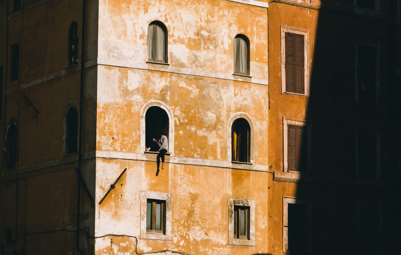Person hanging put from an arched window of a antique and discolored yellow building with shades in Rome Lazio in Italy