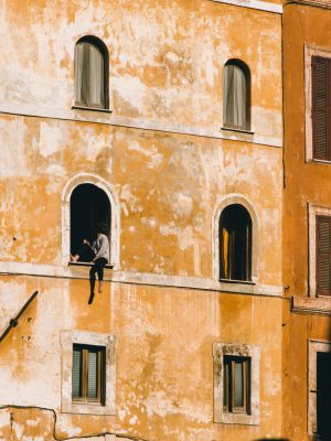 Person hanging put from an arched window of a antique and discolored yellow building with shades in Rome Lazio in Italy
