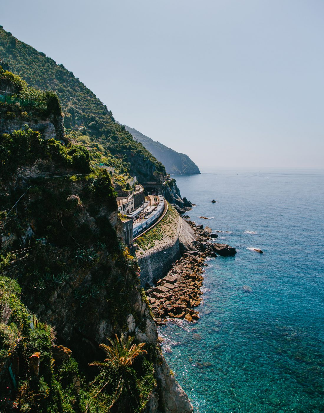 Train station in the middle of a hill with trains crossing over a rocky beach and crystal blue sea surrounded by mountains and plants in Liguria Italy