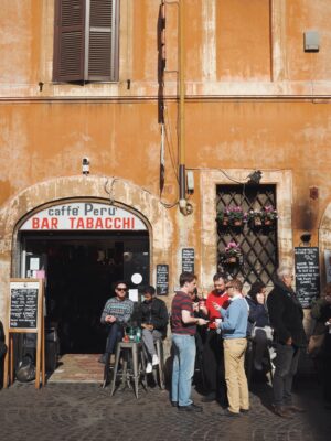 Caffè Perù" - La gente si riunisce fuori da un edificio color arancio con un cartello "Caffè Perù Bar Tabacchi", che incarna un bar tradizionale italiano. Alcuni siedono a un tavolino, mentre altri stanno in piedi a chiacchierare. Menù e fiori in vaso decorano l'area, mentre la luce del sole proietta ombre sulla strada acciottolata.