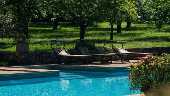 Una piscina azzurra e trasparente con sedie a sdraio al Borgo della Marmotta, circondata da alberi ed erba verde, con la luce del sole che filtra tra le foglie.