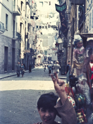 [Image Title] - A bustling, narrow street captures the essence of Italian lifestyle with laundry hanging overhead. People wander in the distance, and a boy playfully holds up two fingers. Quaint shops line the street, a PANTOFOLE sign visible, all contributing to the images vintage, lively feel.