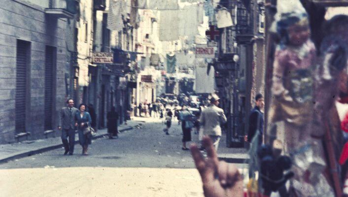 [Image Title] - A bustling, narrow street captures the essence of Italian lifestyle with laundry hanging overhead. People wander in the distance, and a boy playfully holds up two fingers. Quaint shops line the street, a PANTOFOLE sign visible, all contributing to the images vintage, lively feel.