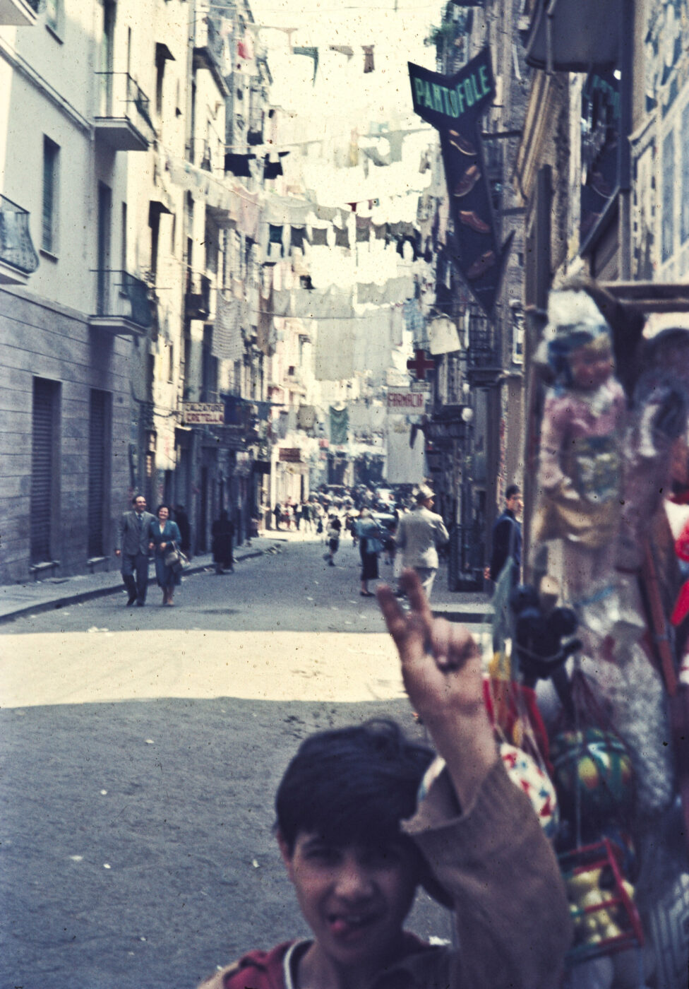 [Image Title] - A bustling, narrow street captures the essence of Italian lifestyle with laundry hanging overhead. People wander in the distance, and a boy playfully holds up two fingers. Quaint shops line the street, a PANTOFOLE sign visible, all contributing to the images vintage, lively feel.