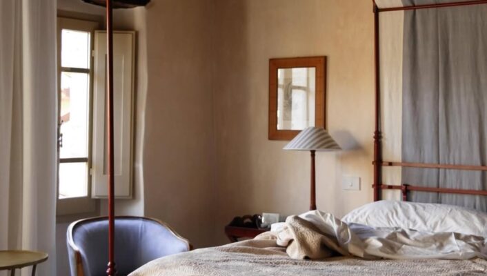 Un'accogliente camera da letto al Borgo della Marmotta con letto a baldacchino rosso, morbida biancheria da letto, lampada, poltrona, tavolo, finestra e pareti beige.