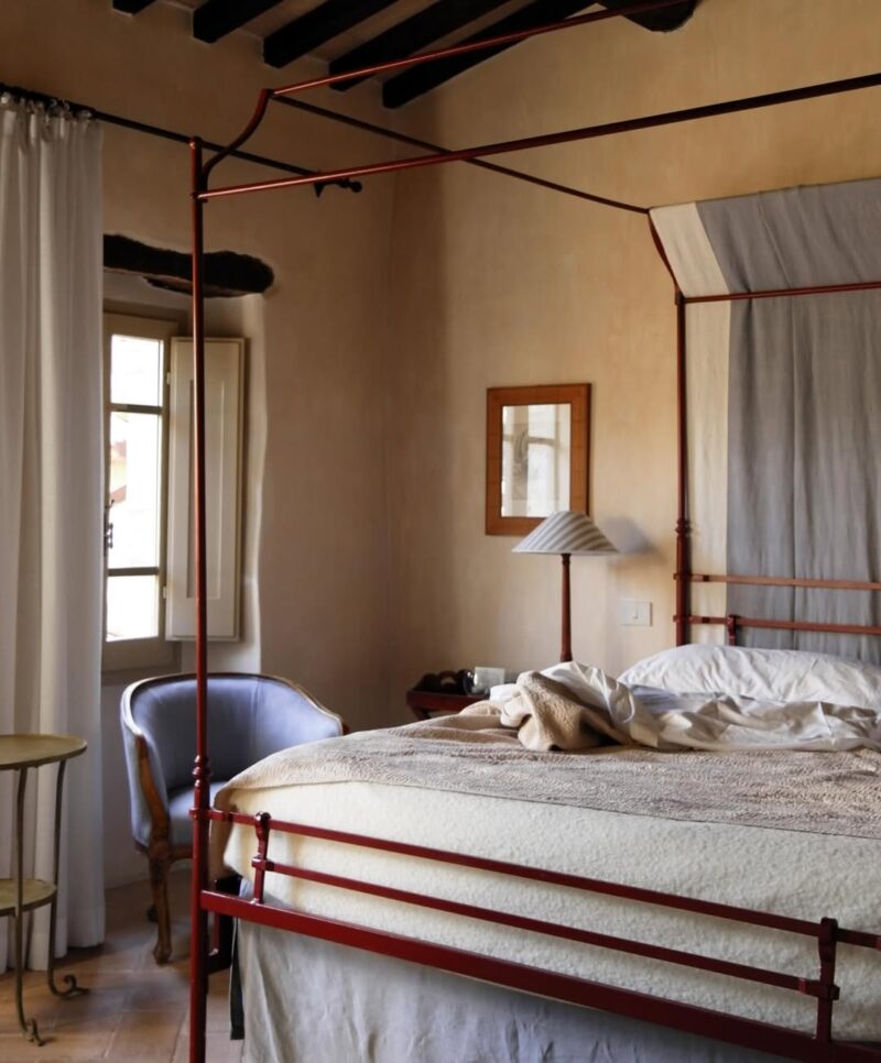 A cozy bedroom at Borgo della Marmotta: red canopy bed, beige bedding, blue armchair, side table, lamp, light curtains, sunlit.