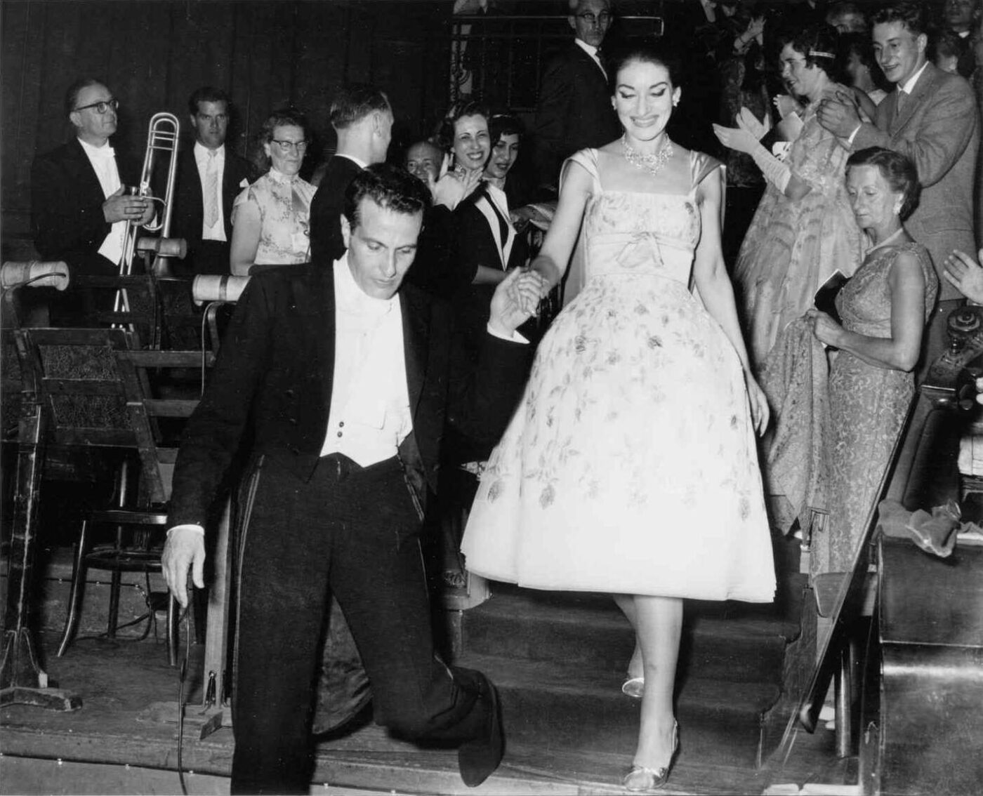 An Elegant Evening at the Theatre - Una donna in un elegante abito italiano viene scortata giù per le scale da un uomo in abito formale. Sono circondati da persone che applaudono, con un musicista che tiene un trombone sullo sfondo. La scena si svolge come una di quelle serate magiche con nomi meno noti che onorano il teatro.
