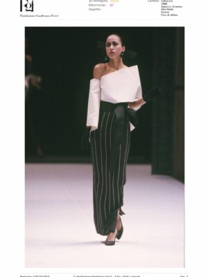 Italian Fashion Spotlight - Model walking down the runway wearing a stylish outfit with an off-shoulder white top and a black and white vertical striped skirt featuring a wide black belt. The ensemble, reminiscent of lesser-known Italian fashion names, is accessorized with black gloves and high-heeled shoes.