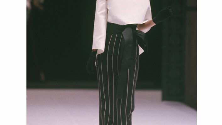 Italian Fashion Spotlight - Model walking down the runway wearing a stylish outfit with an off-shoulder white top and a black and white vertical striped skirt featuring a wide black belt. The ensemble, reminiscent of lesser-known Italian fashion names, is accessorized with black gloves and high-heeled shoes.