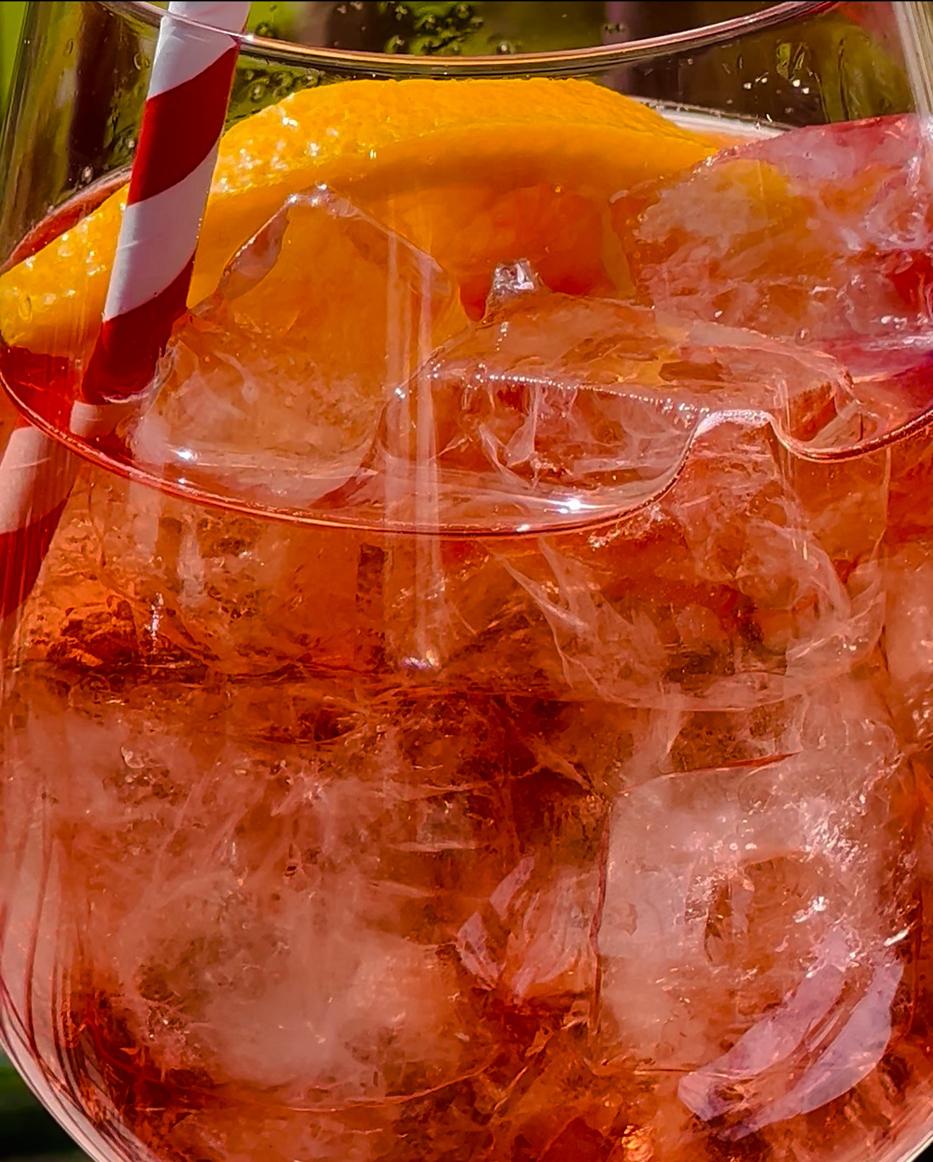 Aperol Spritz - A close-up of a glass filled with ice, an orange slice, and a red-and-white striped straw, featuring a vibrant reddish-pink Aperol Spritz.