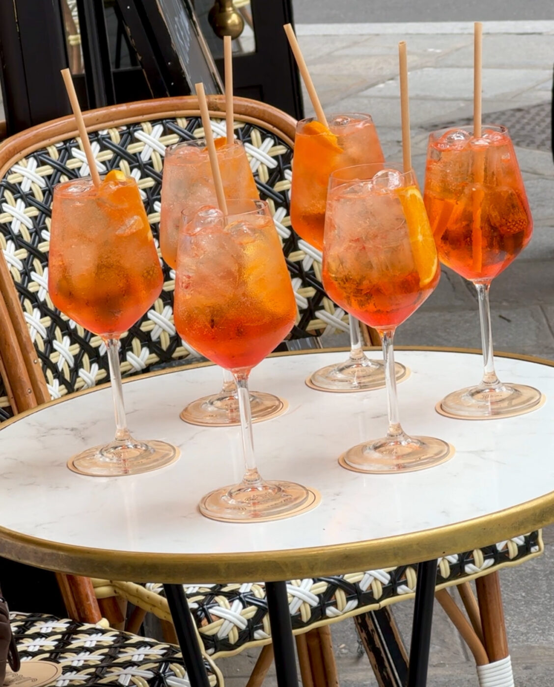Six glasses of Aperol Spritz cocktails with ice and orange slices sit on a round white marble table, each with a straw. The table is at an outdoor café with patterned chairs visible.