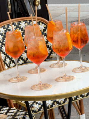 Six glasses of Aperol Spritz cocktails with ice and orange slices sit on a round white marble table, each with a straw. The table is at an outdoor café with patterned chairs visible.