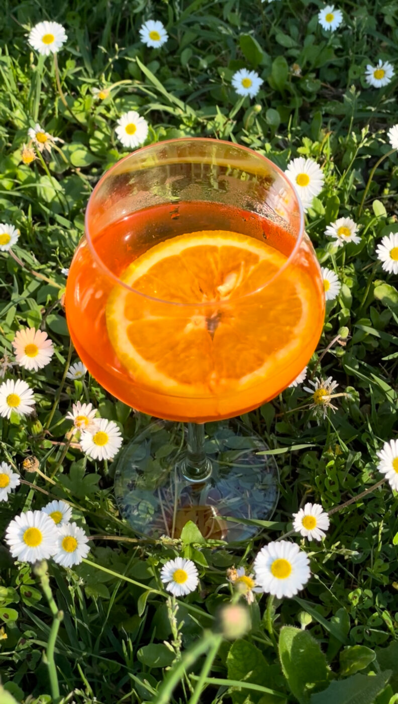 A glass of orange-colored Aperol Spritz with a slice of orange floats inside, placed on green grass surrounded by small white daisies in sunlight.