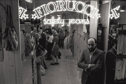 FIORUCCI Safety Jeans - Un uomo in giacca e cravatta è in piedi davanti allinterno di un negozio con pareti in legno, che ricordano le boutique di moda italiane meno note. I clienti curiosano tra i vestiti sugli scaffali. Uninsegna al neon sopra recita FIORUCCI safety jeans, aggiungendo un tocco di estetica vintage e monocromatica.