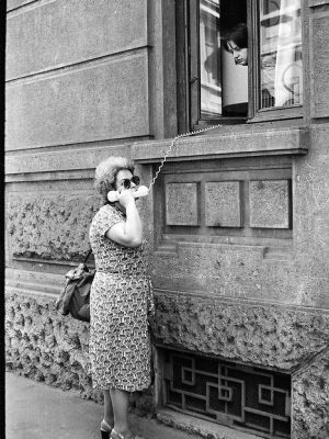 a black and white photo of an older woman standing on the street talking on a phone with the cord running out of an open window