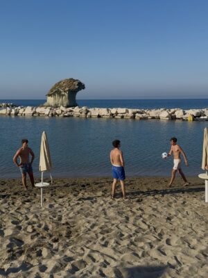 Ischia Island Beach Soccer - Three boys in swim trunks play with a soccer ball on a sandy beach near the water on Ischia Island, Campania. Closed umbrellas and lounge chairs are nearby, as a unique rock formation rises from the sea beneath the clear blue sky.
