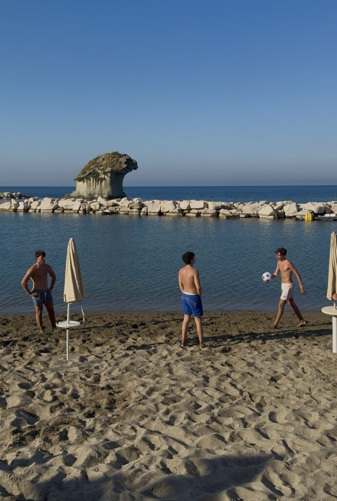 Isola d'Ischia, Campania, scena di spiaggia: tre ragazzi in costume da bagno giocano con un pallone da calcio sulla spiaggia sabbiosa dell'Isola d'Ischia, vicino all'acqua calma; sullo sfondo sono visibili ombrelloni chiusi e una formazione rocciosa sotto un cielo azzurro e limpido.