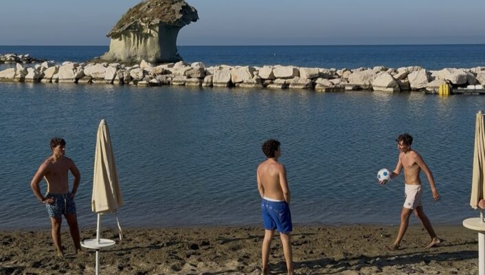 Isola d'Ischia, Campania, scena di spiaggia: tre ragazzi in costume da bagno giocano con un pallone da calcio sulla spiaggia sabbiosa dell'Isola d'Ischia, vicino all'acqua calma; sullo sfondo sono visibili ombrelloni chiusi e una formazione rocciosa sotto un cielo azzurro e limpido.