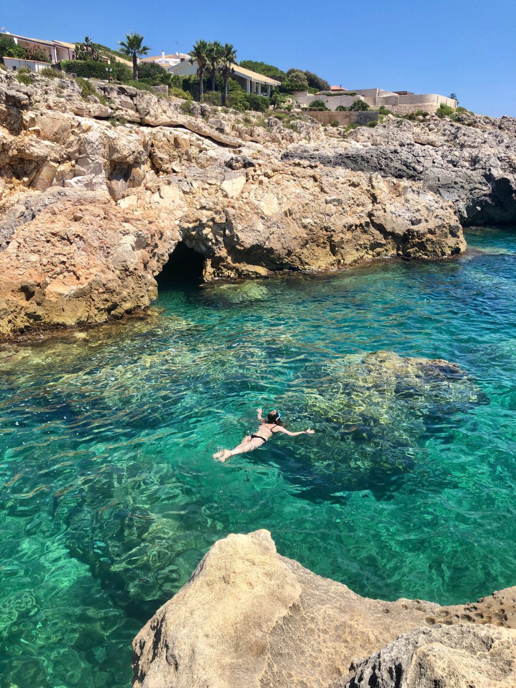 Ortigia - a woman swimming in crystal clear, teal water with sand colored rocks