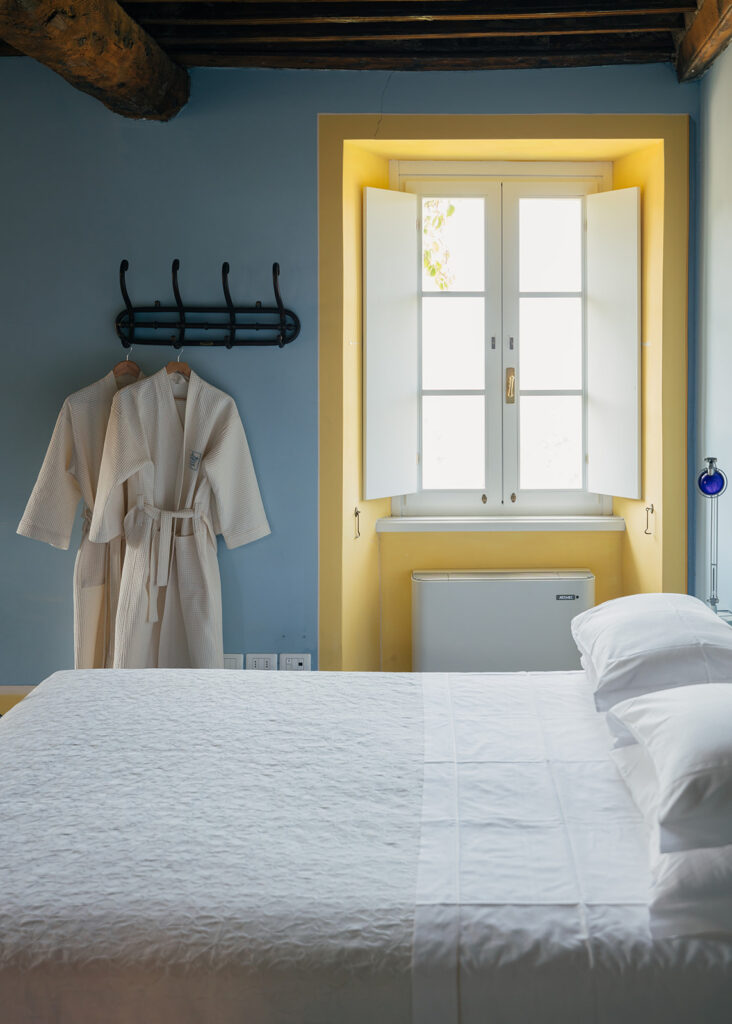 a room at La Sosta di Ottone III with pale blue walls, yellow framing the window, and two white robes hanging