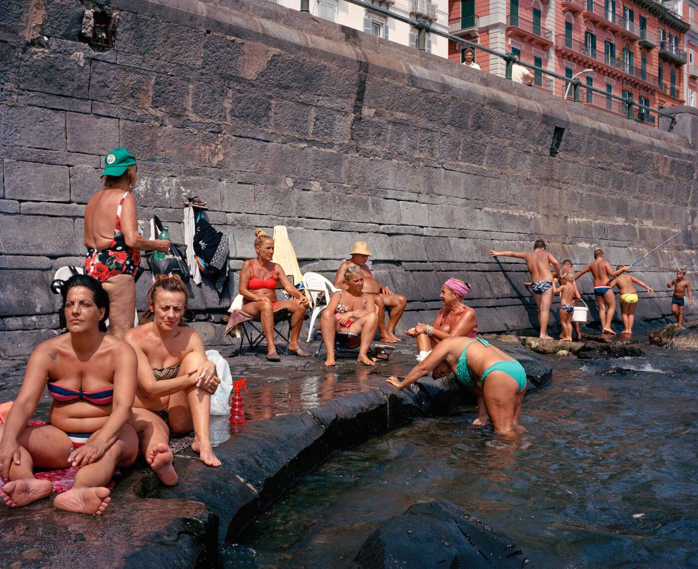 a group of people sitting along the rocky wall against the sea in Naples